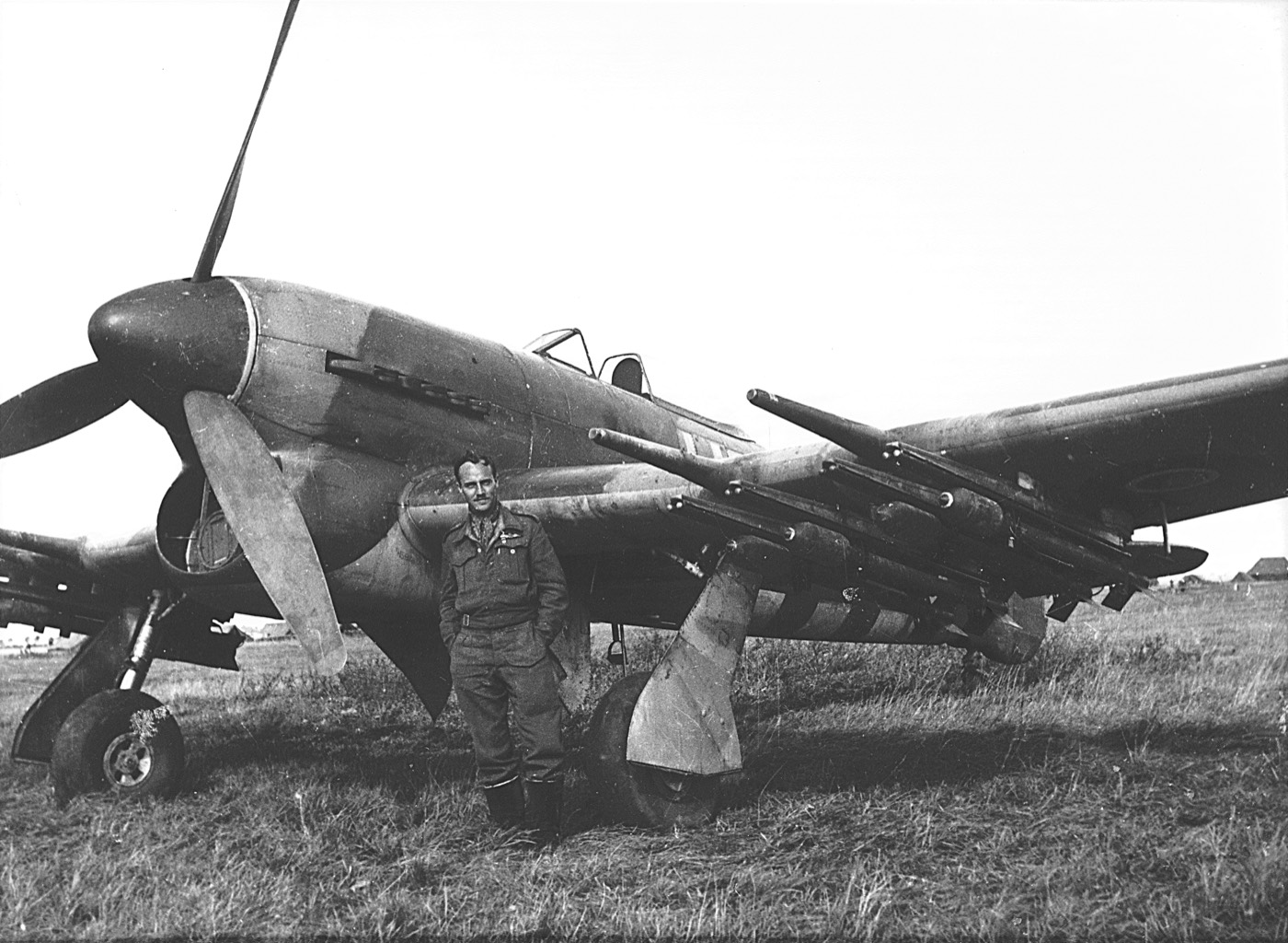 A pilot in battledress standing beside a rocket-armed Hawker Typhoon on a grass airfield